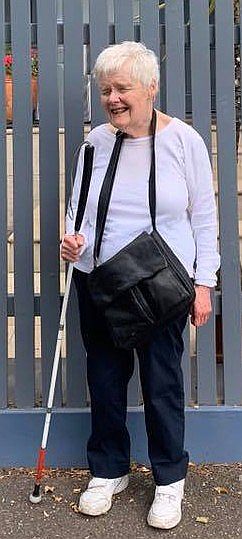 Photo of Mary Wynne standing in front of a blue wooden fence.  Mary has white hair and is wearing a long sleeve light coloured top, blue pants and white shoes.  She has a big black bag around her neck, a white cane in her right hand, and a smile on her face.