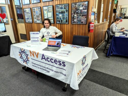 Sean sitting at the NV Access table at SVRC.  The table has an NV Access tablecloth, fliers, business cards and a laptop demonstrating NVDA.