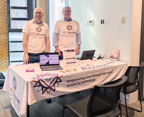 Quentin and James at Tech Fest Adelaide, 2025, in NV Access tshirts at a table with an NV Access banner, brochures, and a computer with NVDA running and the training material on show.