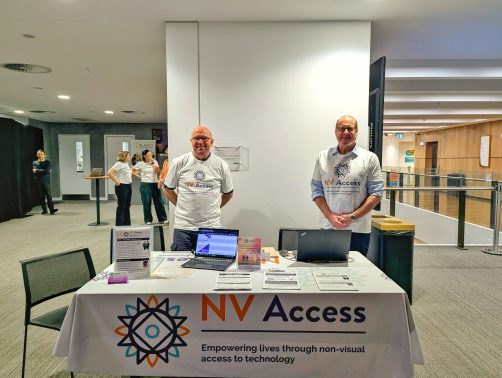 Quentin and James at a table with NV Access table cloth, brochures and laptop